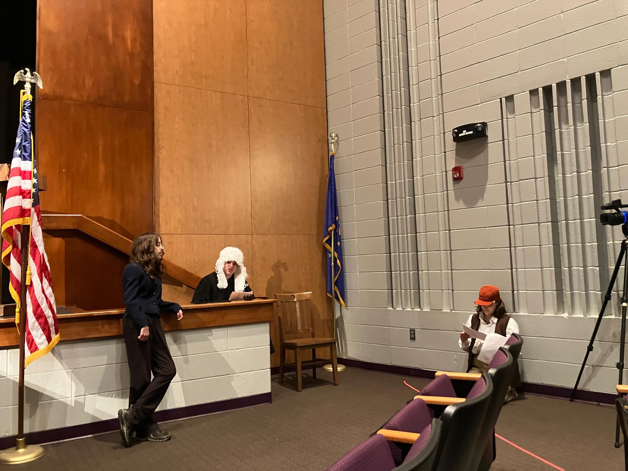Seventh grader Eleanor Bender looks at freshman James Lindsey who acts as a judge. Eighth grader Leslie Stockton sits on the sidelines and reviews the script (Courtesy Photo of Maria Wishart).  